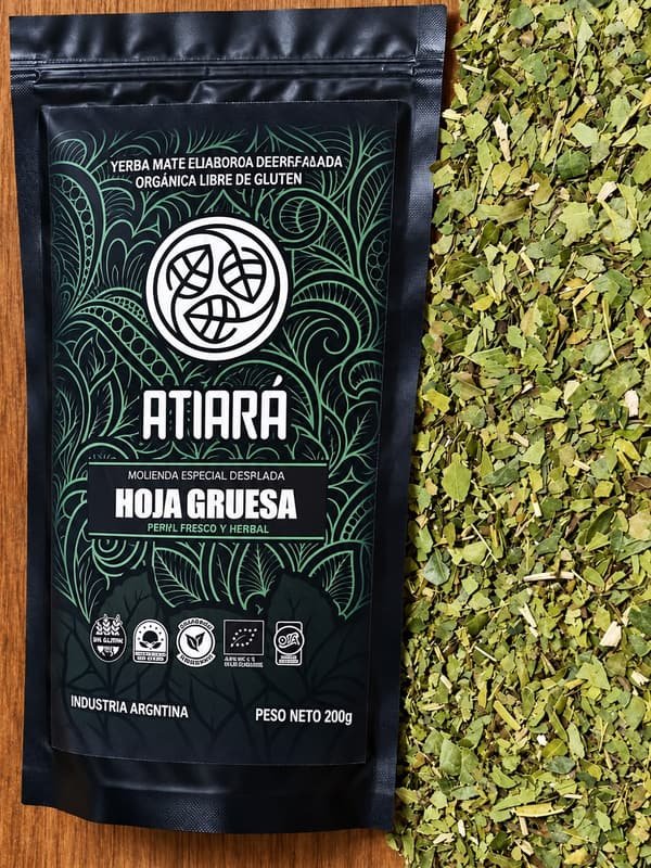 Paquete de Atiará Hoja Gruesa junto a yerba de hojas grandes, sobre una mesa de madera.